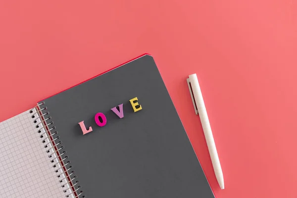 Renkli harflerle yazılmış gri defterli, açık spiral bir defter. Aşkla yazılmış ve pembe arka planda beyaz bir kalem. Sevgililer günü mesajı, aşk ve romantizm konsepti için hazırlan. İleti yaz