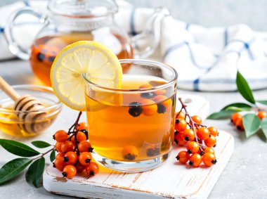 Taze limon dilimi ve parlak böğürtlen dalıyla süslenmiş buharda pişmiş bir deniz dikenli çay. Bu şifalı bitkisel karışım beyaz bir peçeteye ısı, rahatlık ve bağışıklık desteği sağlar..