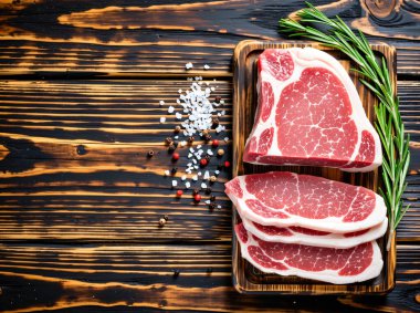 Tahta bir tahtanın üzerinde çiğ biftek, çeşitli baharatlar, sarımsak ve koyu dokulu arka planda biberiye..