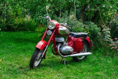 Krom yakıt deposu detayları ve siyah koltuğu olan, yeşil ağaçların ve bahçenin arka planında duran klasik kırmızı retro motorsikletin yan görüntüsü..