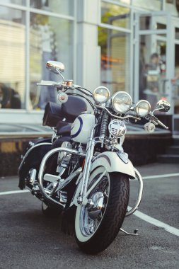 Cam bir binanın önüne park etmiş klasik bir beyaz ve mavi kruvazör motosikletinin dikey portresi. Çekim, krom ön çatal, üçlü farlar, geniş gidonlar ve beyaz duvar lastikleri üzerine odaklanarak klasik bisiklet stilinin özünü yakalıyor..