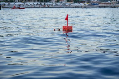 Mavi yüzey üzerinde küçük bir bayrak dalgalanan parlak kırmızı kare şamandıraya odaklanan yatay bir çekim. Plaj şemsiyeli kıyı şeridi yumuşak bokeh arka planda görünür..