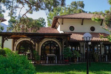Türkiye 'de güzel bir villanın ya da restoran binasının yatay görüntüsü. Kırmızı kil kil kiremit çatı, ahşap terastaki kemerli pencereler ve yemyeşil ağaçlar ve çam ağaçları arasında yuva yapmış yuvarlak dekoratif pencerelere sahiptir..