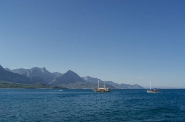 Derin mavi Akdeniz 'de yelken açan iki ahşap turist gemisinin yer aldığı geniş yatay bir deniz manzarası. Arkaplan, berrak bir gökyüzünün altındaki devasa, puslu dağ sıraları tarafından yönetiliyor..