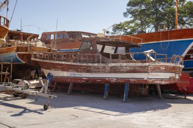 Yıpranmış ahşap motorlu bir teknenin kuru bir rıhtımda geniş yatay görüntüsü. Parlak bir güneşin altında arka planda farklı tamir aşamalarında pek çok tekne görülebilir..