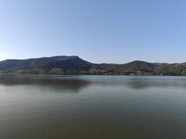 Güneşli bir günde berrak mavi gökyüzünün altındaki uzak dağları yansıtan sakin bir nehir.