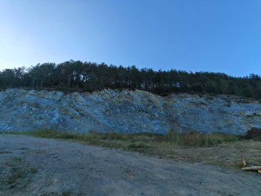 Dik kayalık yamaçlar, açık mavi bir gökyüzünün altında yüksek ağaçlardan oluşan yoğun bir ormanın tepesinde çıplak taşlar ve çakıllar var.