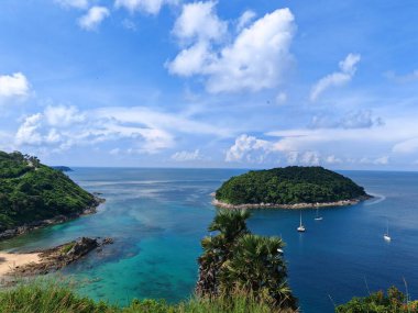 İki küçük yeşil ada, turkuaz su ve parlak bir gökyüzü altında yüzen teknelerin bulunduğu tropik bir körfezin manzarası.