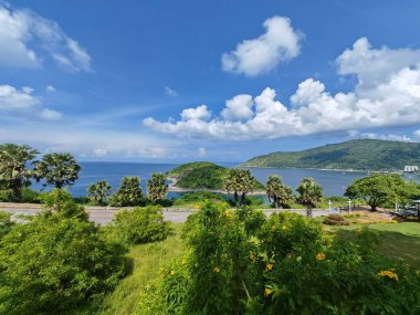 Bereketli yeşillik, palmiye ağaçları, beyaz bulutlu mavi bir gökyüzü ve arka planda dağlar olan güzel bir tropikal kıyı manzarası.