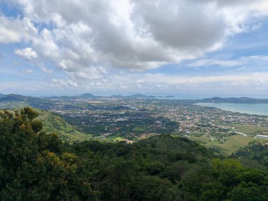 Yeşil tepeler ve bulutlu mavi gökyüzü ile çevrili, bir şehir ve kıyı şeridi manzarası
