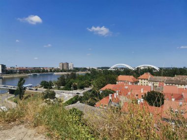 Panoramik şehir manzarası bir nehir, modern köprü, kırmızı çatıları olan konut binaları ve açık mavi gökyüzünü gösteriyor.