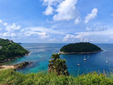 Berrak turkuaz suyu, yemyeşil adaları ve parlak gökyüzü altında yelkenlileri olan sakin bir tropikal koy manzarası.