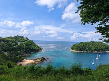 Turkuaz su, kumlu plaj, demirli yelkenliler ve bulutlu, parlak mavi gökyüzünün altında yemyeşil tepeler olan sakin bir koy.