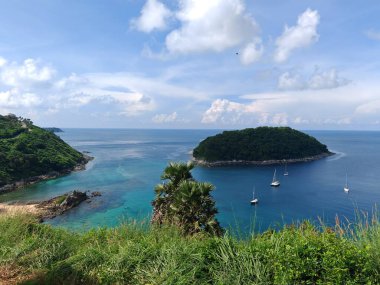 Açık mavi denizle çevrili küçük tropikal ada parçalı bulutlu bir gökyüzünün altında kıyıya vuran yatları var.