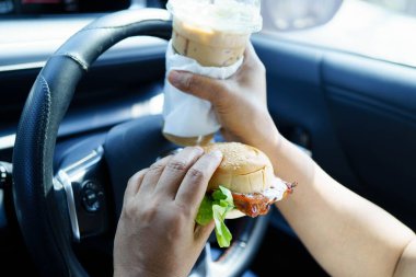 Asyalı kadın sürücü arabada hamburger yiyerek bekliyor..