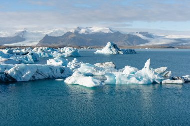 Derin mavi sularda yüzen buzdağlarının manzarası. Bulutlu gökyüzünün altında karla kaplı dağlar..