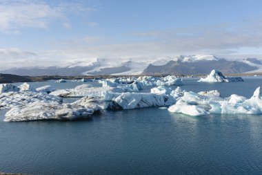 Yüzen buzdağları, arka planda karla kaplı dağlarla buzul gölünün sakin yüzeyinde huzur içinde sürükleniyor..