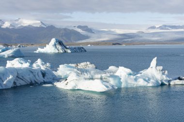 Sakin bir gölde sürüklenen buzdağlarının huzurlu görüntüsü. Arkaplanda karla kaplı dağlar yükselir, ve çok güzel bir zıtlık yaratır..