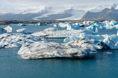 Buzdağları sakin mavi sularda yüzer. Arka planda karla kaplı dağlar, parlak güneş ışığı.. 