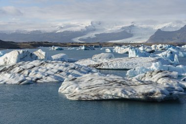 Uzaklarda karla kaplı dağlarla mavi bir buzulda yüzen büyük buzdağlarının manzarası..