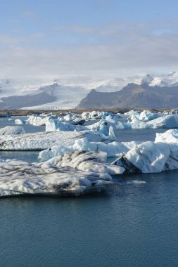 Parçalı bulutlu bir gökyüzünün altında karla kaplı dağların desteklediği buzdağlarının buzdağlarında yüzdüğü sakin bir manzara..
