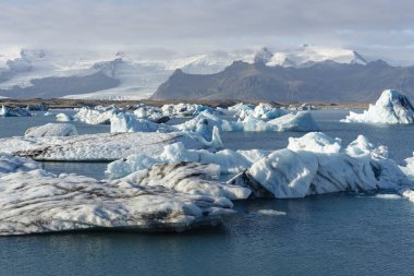 Uzaktaki karlı dağlarla birlikte buzul gölünde huzur içinde yüzen tüm şekil ve boyutlardaki buzdağlarının nefes kesici görüntüsü..