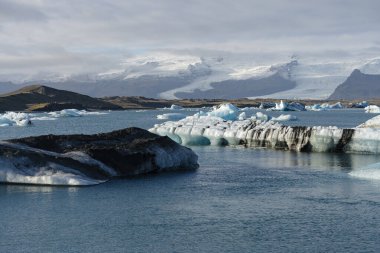 Buzdağları, bulutlu bir gökyüzünün altındaki karlı dağların arka planına doğru sakin bir şekilde sürüklenir..