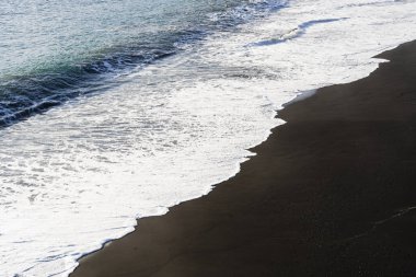Güneşli bir günde kıyıya vuran siyah kum ve köpüklü beyaz dalgalar arasındaki güzel zıtlık.