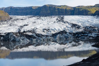 Uzakta dağlık arazisi olan büyük bir buzulun önünde yansıtıcı bir su kütlesi duruyor..