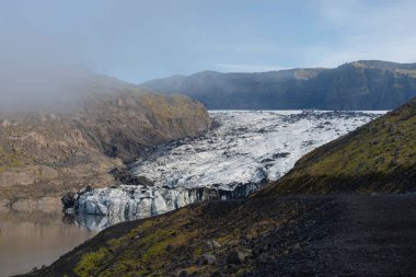 Kayalık yamaçlar arasında bir buzul, sisli dağların arkasına kurulmuş sakin ve yansıtıcı bir göle iniyor..