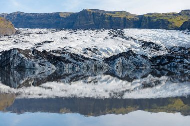 Buzul oluşumları berrak göle yansıyor. Arkasında mavi gökyüzü olan kayalık tepeler var..