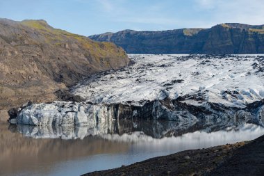 Buzul dilinin sakin bir gölle buluşmasını, berrak sulardaki dağları yansıtan manzaralı bir manzara..