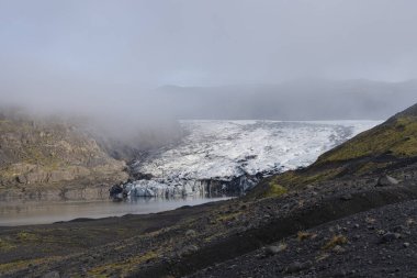 Buzulun güzel manzarası, buzulun etrafını saran dağlar. Ön planda volkanik kayalar görünüyor..