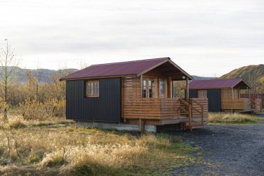 Çimenli bir arazide birbirine benzeyen iki kulübe var. Ahşap verandaları ve karanlık duvarları olan kırmızımsı bir çatıları var..