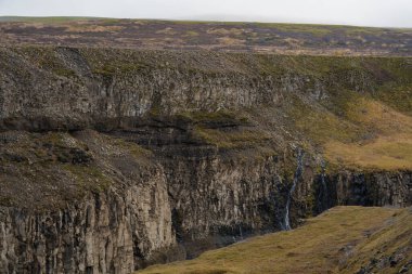 Yüzeyinden dökülen küçük, narin bir şelale ile engebeli bir uçurum manzarası. Gökyüzü bulutlu.