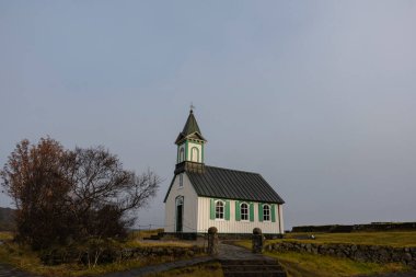 Çan kulesi olan küçük, beyaz bir kilise bulutlu bir gökyüzünün altında çimenli bir tarlada duruyor. Taş yol kapılarına kadar uzanıyor..