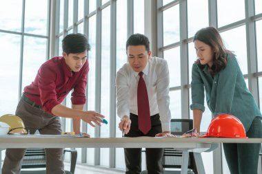 Üç inşaat mühendisi ve mimar toplantı masasında mühendis şapkasıyla çalışma masasında beyin fırtınası yapıyor.