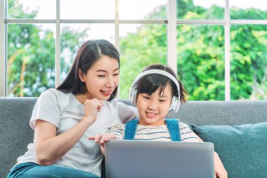 Asyalı anne ve kızı film izlerken eğleniyorlar ve evdeki koltukta dizüstü bilgisayardan müzik dinliyorlar..