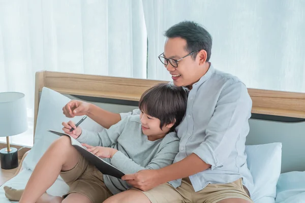 Father and son is sitting on the bed doing homework writing on a book together, Father teaching son to write with smile.