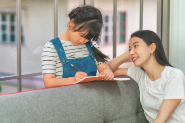 Asyalı Anne Öğretmen, evdeki oturma odasında kızını ödev yaparken izliyor. Aile dayanışması kavramı için.
