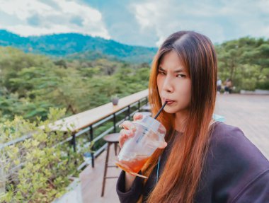 Komik yüzlü güzel Asyalı kadın buzlu amerikano kahvesi içerken orman ve dağ doğası geçmişi olan bir kafede oturuyor.