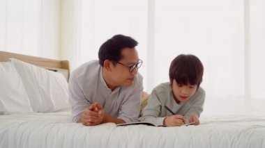 Asyalı baba, oğlunun okul ödevlerine evde bir yatakta yardım ediyor..