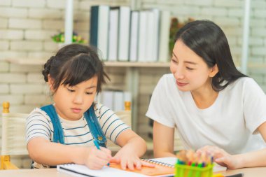 Anne Öğretmen, kızına, evdeki oturma odasındaki kanepenin boyama kitabında ödev yapmayı öğretiyor. Aile dayanışması kavramı için.