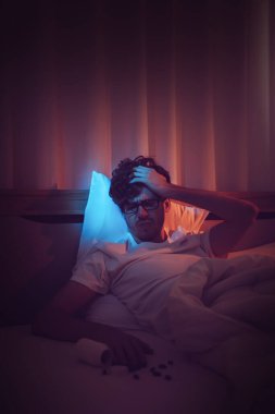 Depresyondaki uykusuzluk çeken beyaz adam gece geç saatlerde stres ve intihar konsepti için siyah haplarla başını yatakta tutuyor..