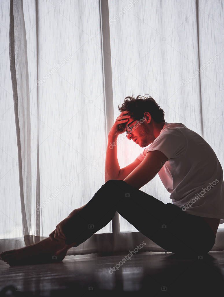 Caucasian male is sitting on the floor by the windows with depressed emotion over trouble and problem.