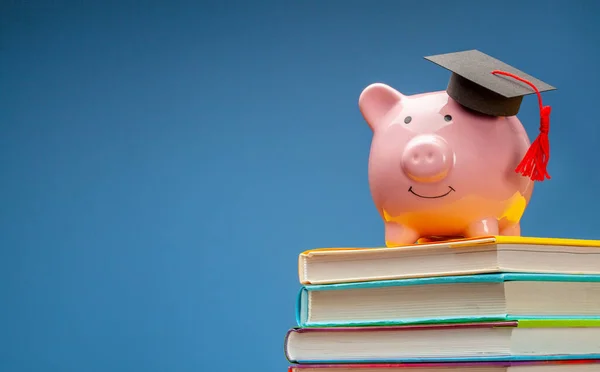 Sparschwein mit Diplom-Hut auf Bücherstapel. Blauer Hintergrund. Kopierraum für Text — Stockfoto