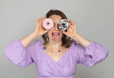 Kız gözlük olarak donut tutuyor ve gri arka planda mor elbisesiyle dilini gösteriyor.