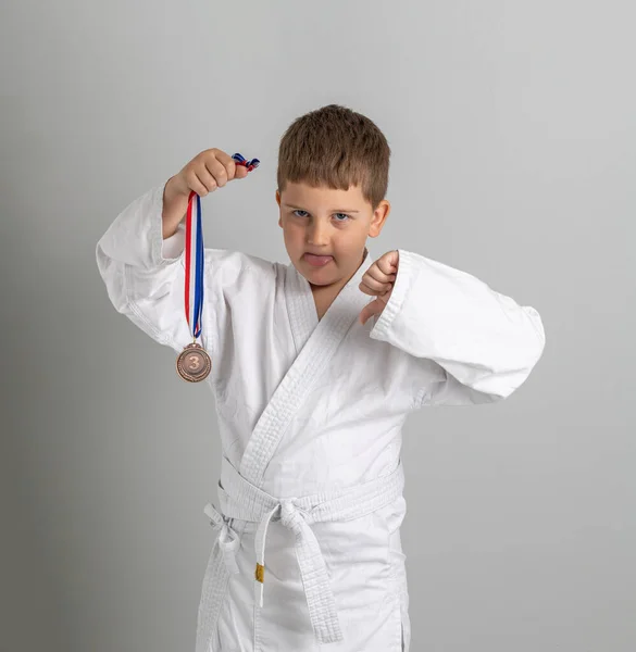 Chłopiec w kimono ma brązowy medal za trzecie miejsce w konkursie. Nieszczęśliwe dziecko pokazuje język i kciuki w dół. Szare tło — Zdjęcie stockowe