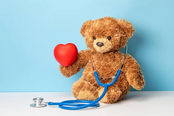 Médico de niños o médico de familia. El oso de peluche con un estetoscopio sostiene el corazón rojo — Foto de Stock