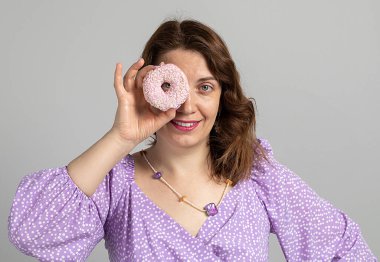 Kızın elinde gözlük gibi bir çörek var. Gri arka planda mor bir elbiseyle gülümseyen kız.